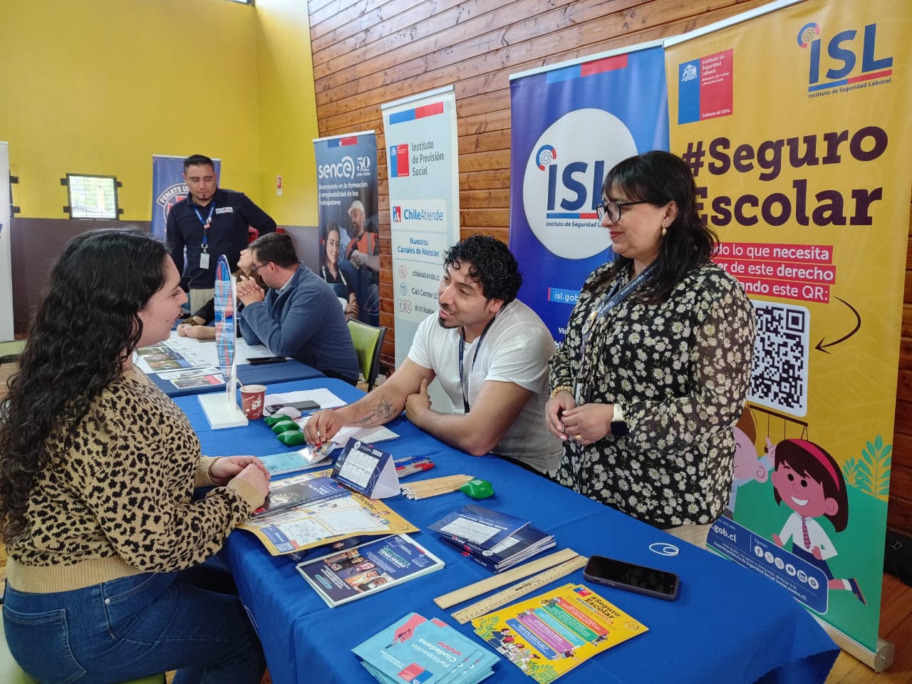 Personas conversan en un stand del ISL, donde se entrega información sobre el Seguro Escolar y se exhiben materiales informativos junto a pendones institucionales.